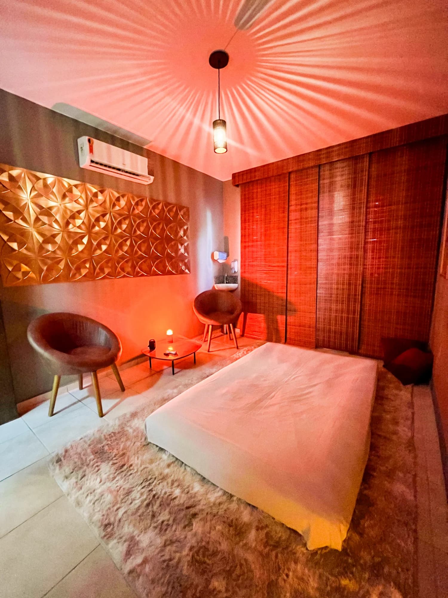 Sala Sândalo do Espaço Cor em luz quente: futon no chão sobre tapete felpudo, painel 3D dourado e persianas de bambu